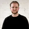 Portrait of a bearded man in glasses, black top, standing against a light brick wall.